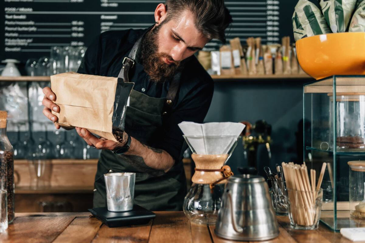 Close up of a young male barista pouring coffee beans into a glass and measure it on digital scale in coffee shop.