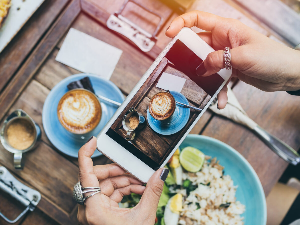 Woman's hands taking photo of coffee cup, Piccolo Latte, brownies with breakfast on wooden table by smartphone, top view.