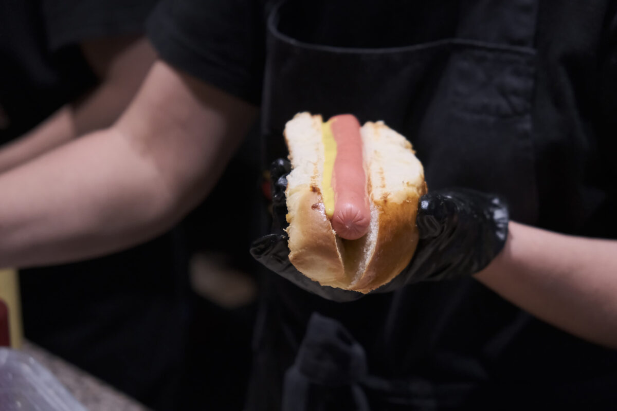 Fresh hot dog with grilled sausage, served outdoors. Cookout american bbq food. Hot-dog with meat and vegetables salad filling closeup in chef's hands. Street food, fast food.