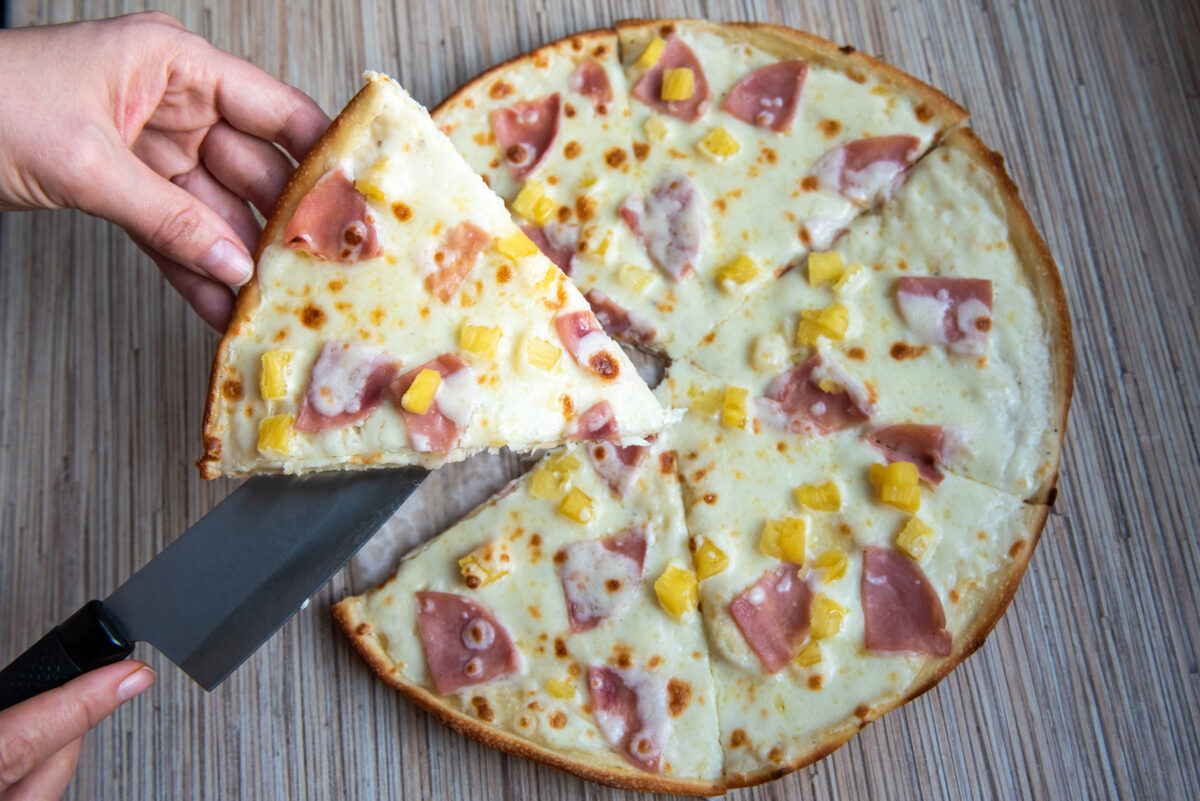 A female hand takes a slice of pizza with a kitchen knife. Italian hawaiian pizza on a wooden table.