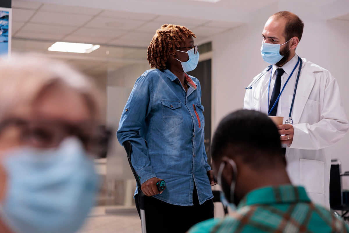 African american patient with crutches in hospital lobby, talking to male doctor about healthcare and recovery. Woman with chronic disability and impairment doing checkup visit.