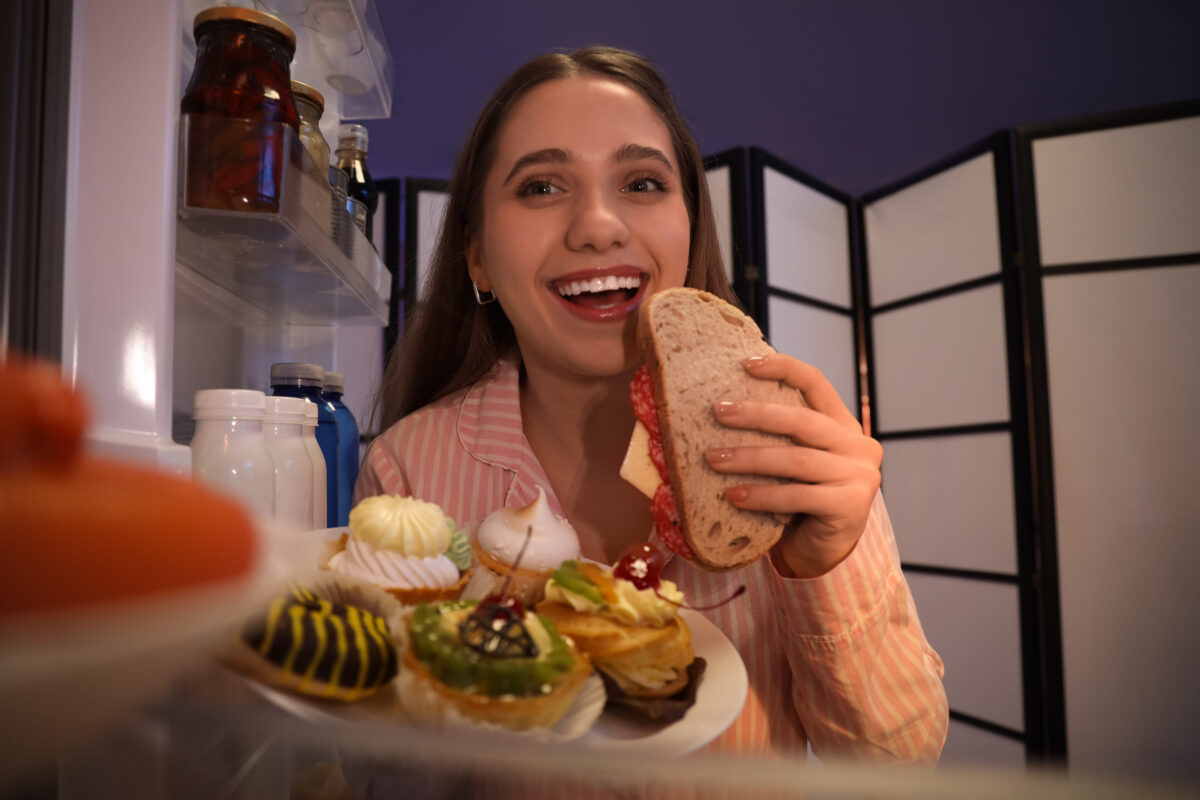 Hungry young woman eating sandwich in kitchen at night, view from inside of refrigerator