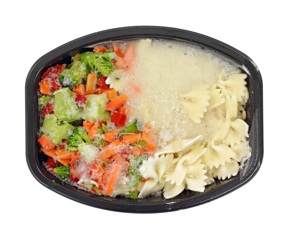 A still frozen TV dinner with pasta and vegetables in black dish.