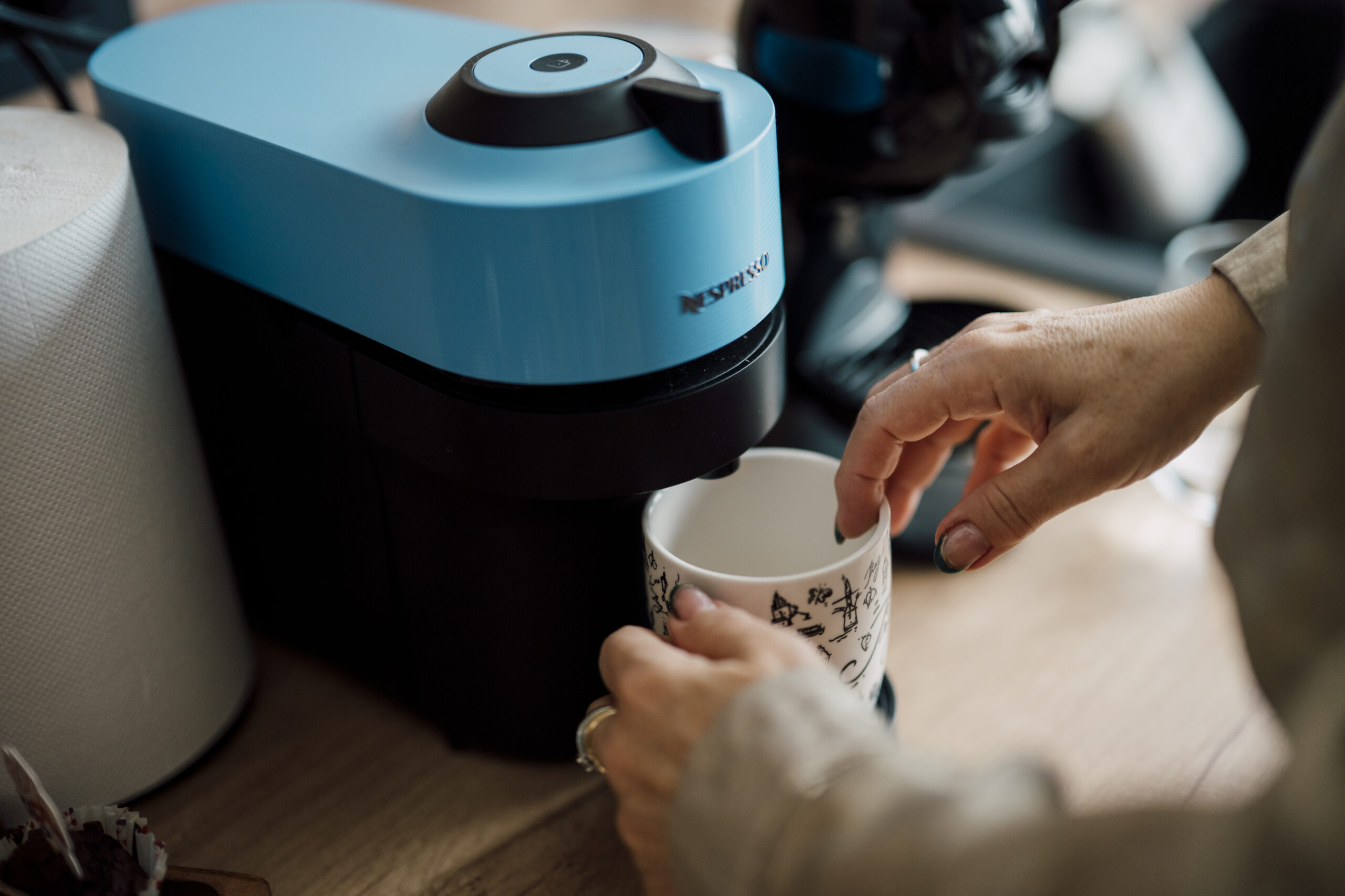 A person prepares coffee using a coffee maker in a kitchen. The machine is blue and the coffee is being poured into a cup. Morning light is present in the room.