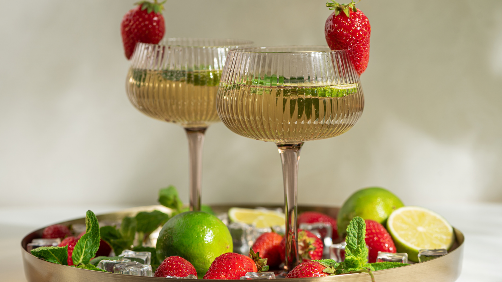 Two sparkling strawberry lime mojito mocktails garnished with fresh mint leaves and whole strawberries, surrounded by limes, strawberries, and ice cubes on a modern golden tray