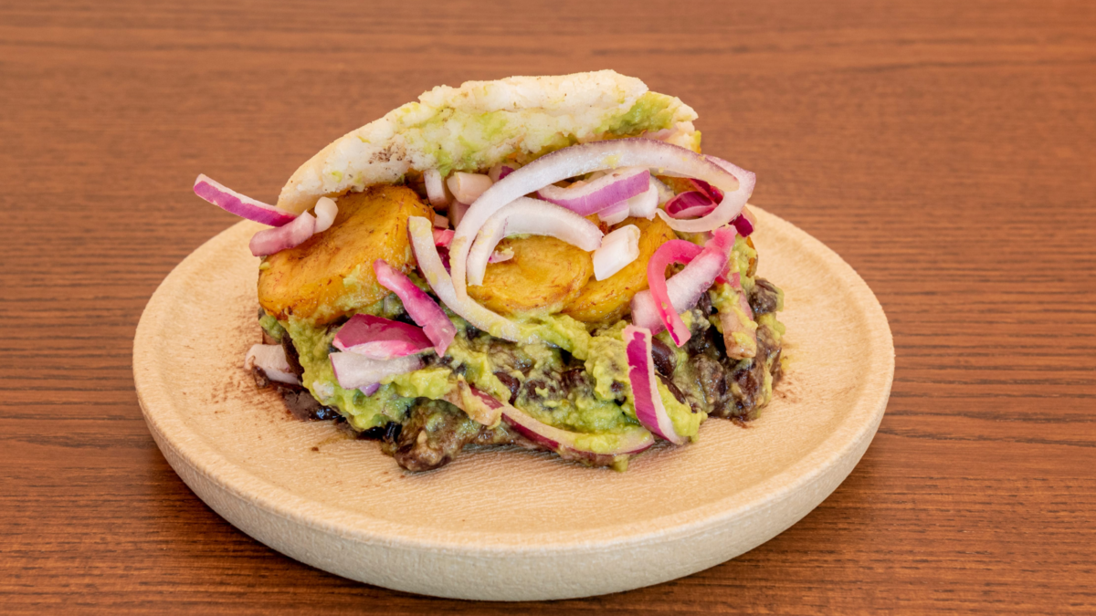 Vegan arepa stuffed to overflowing with lots of red onion, lettuce, guacamole and male banana slices on brown table