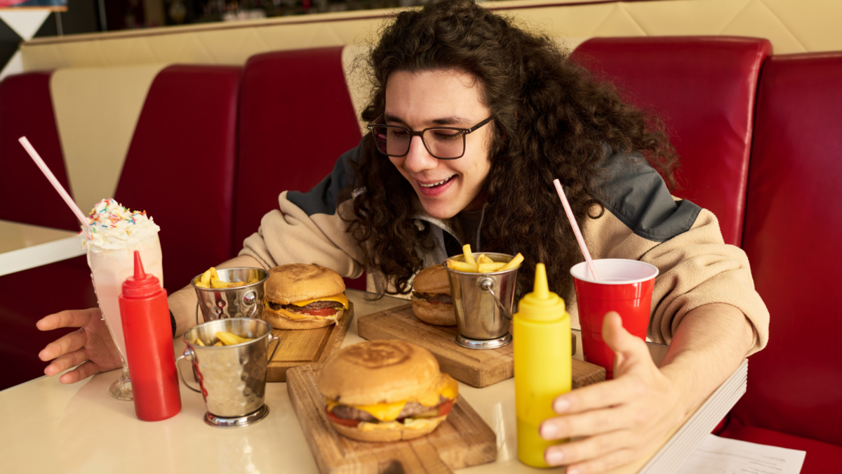 Young Caucasian man with long curly hair smiling and reaching toward cheeseburgers and fries on table in diner, milkshake and soft drink with straws visible nearby