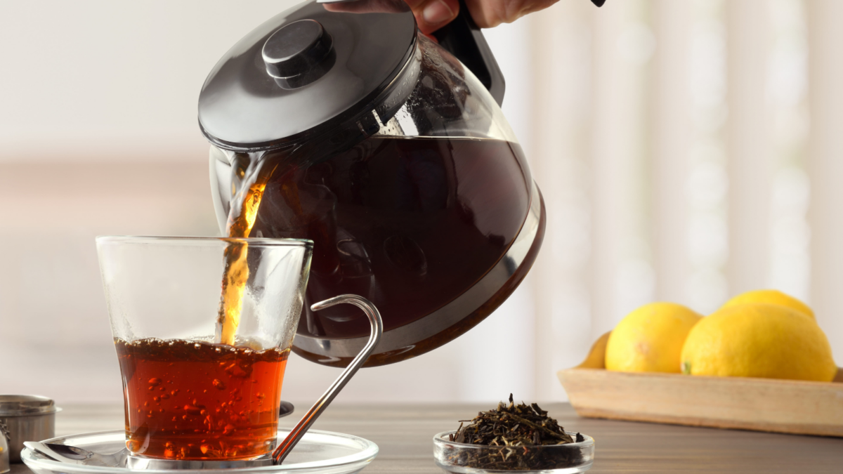 A person is pouring tea into a glass cup on a wooden table. There is a bowl of tea leaves and lemons on the table. Image Credit: davizro/ 123RF 