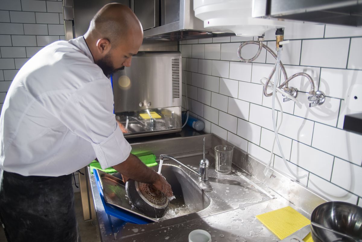 8 Restaurant Secrets That Make Professional Kitchens Run Better Than Most Home Cooks Realize 2 A chef cleans kitchen utensils in a stainless steel sink, maintaining hygiene standards.