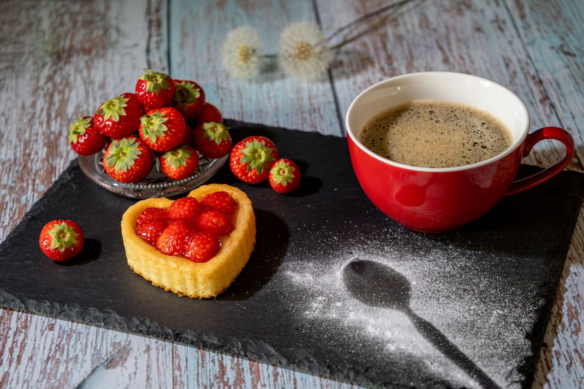 A delightful heart-shaped strawberry tart next to a cup of coffee on a rustic slate board.