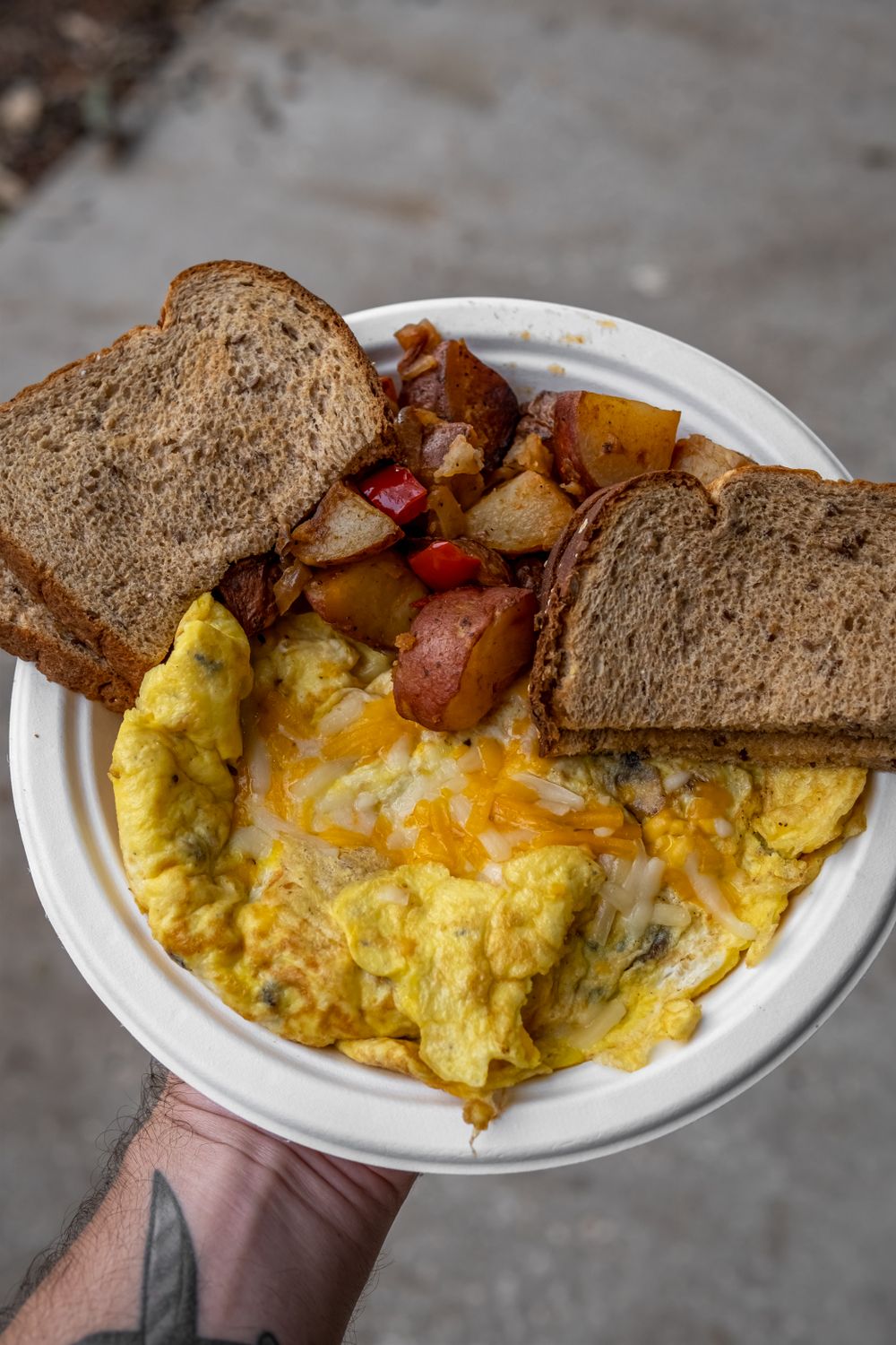 A hearty breakfast plate with scrambled eggs, toast, and potatoes held outdoors.