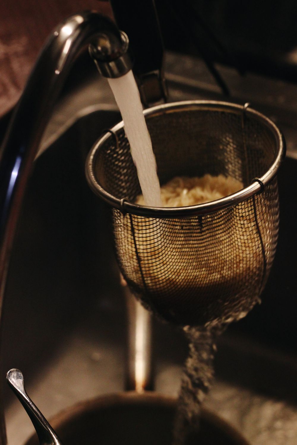 A kitchen scene showing water pouring over noodles in a mesh sieve in a sink.