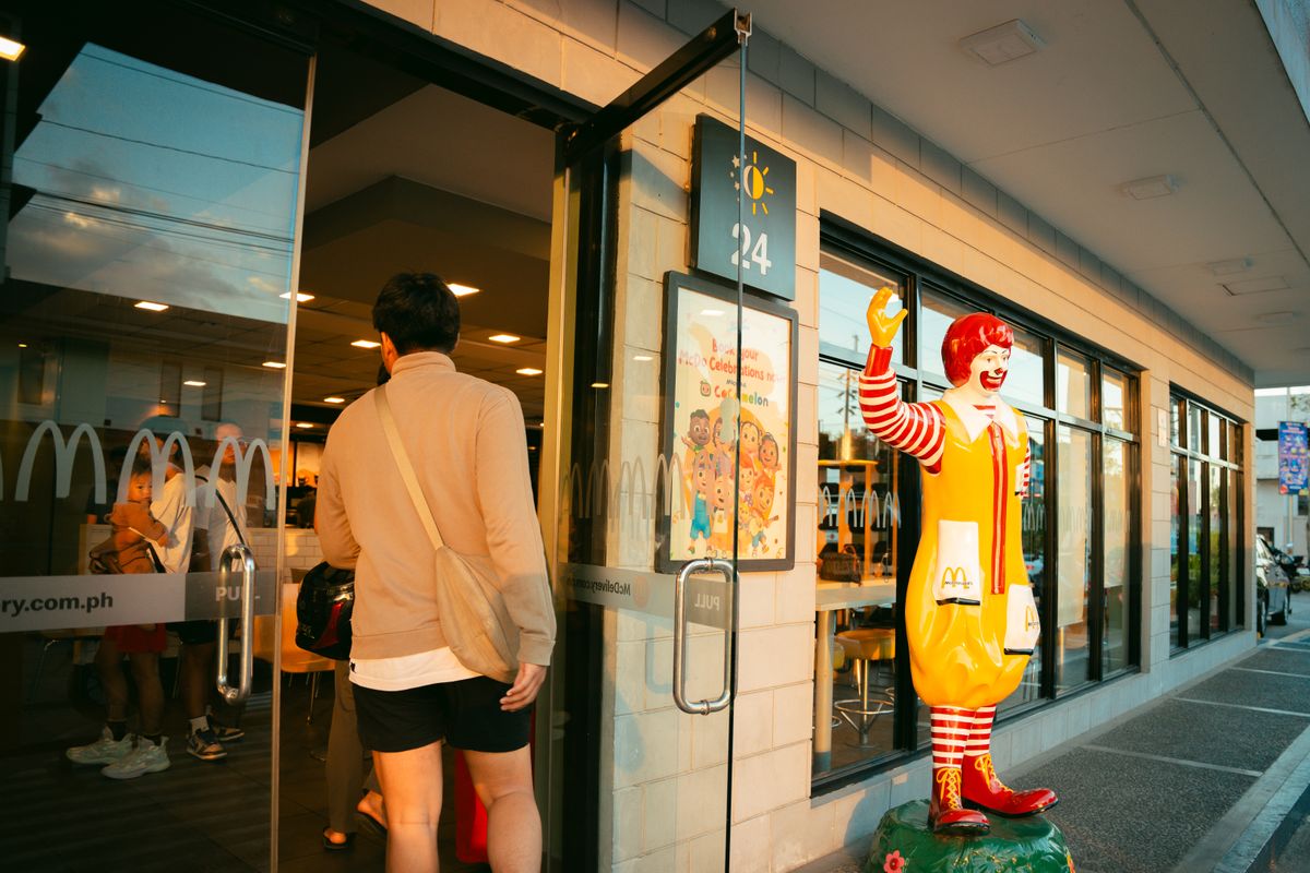 A man in casual attire enters a McDonald's restaurant, past a Ronald McDonald statue, during daylight.