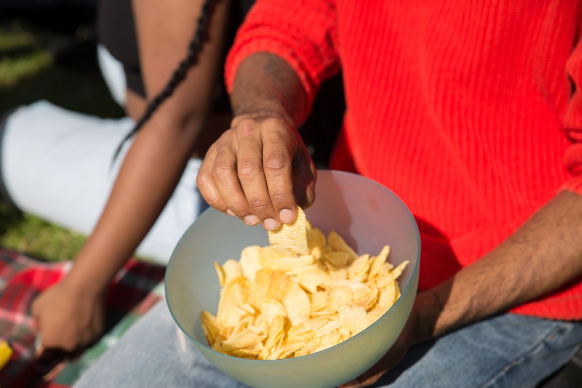 A person in a red sweater holds a bowl of potato chips during a sunny outdoor picnic.