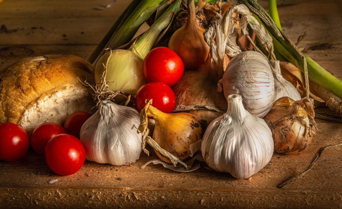 A rustic still life featuring garlic, onions, cherry tomatoes, and sourdough bread.