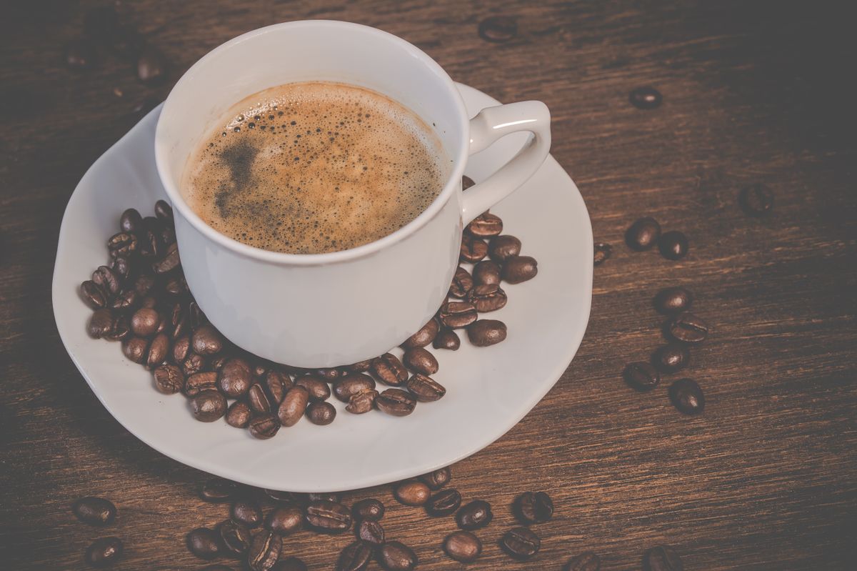A steaming espresso served in a white cup surrounded by roasted coffee beans on a wooden table.