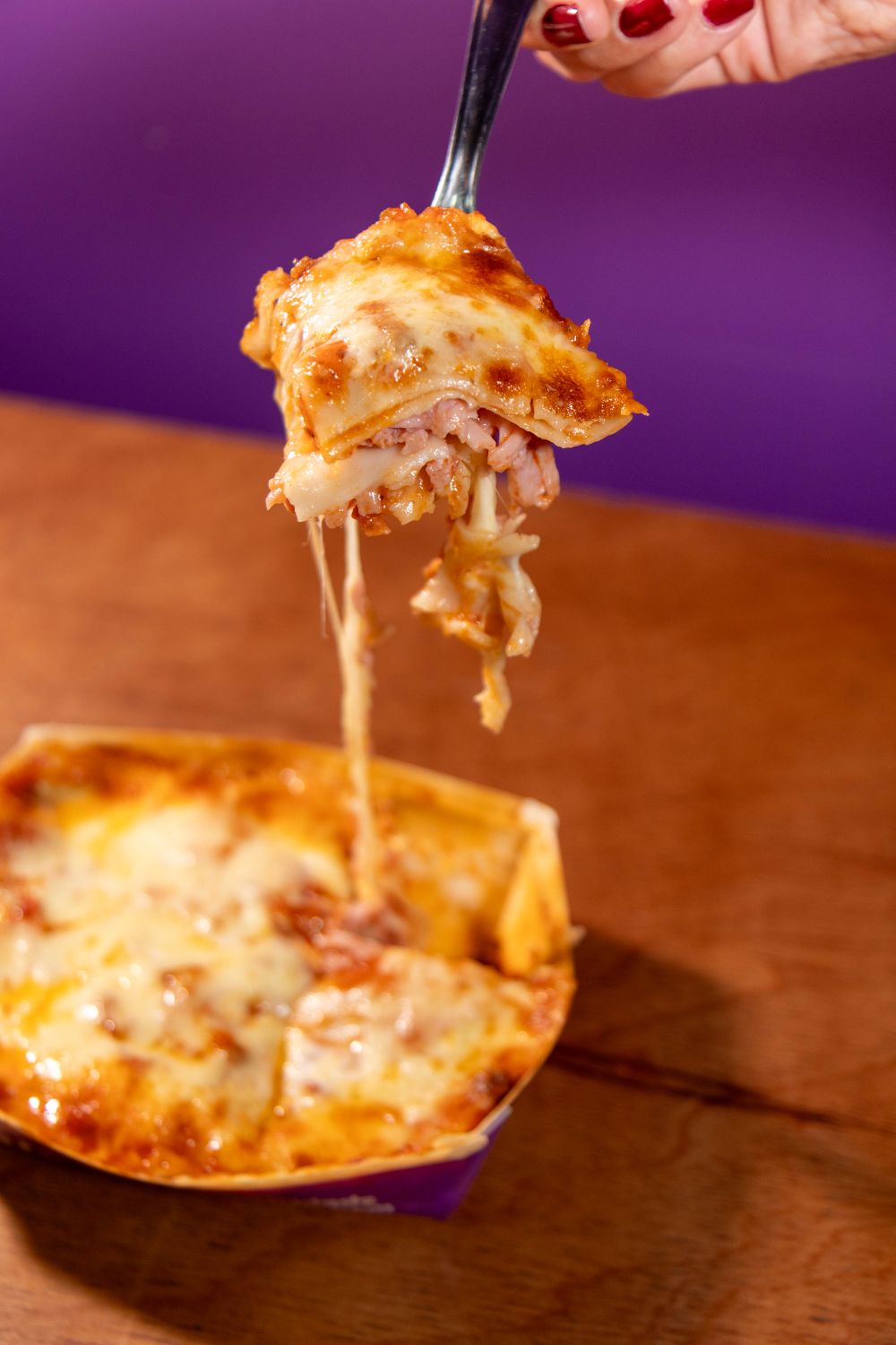 Close-up of a fork lifting a cheesy lasagna slice from a dish, against a vibrant purple background.