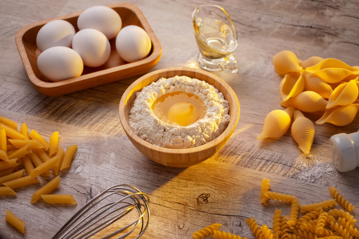 Ingredients for making homemade pasta including eggs, flour, and various pasta shapes, arranged on a kitchen table.