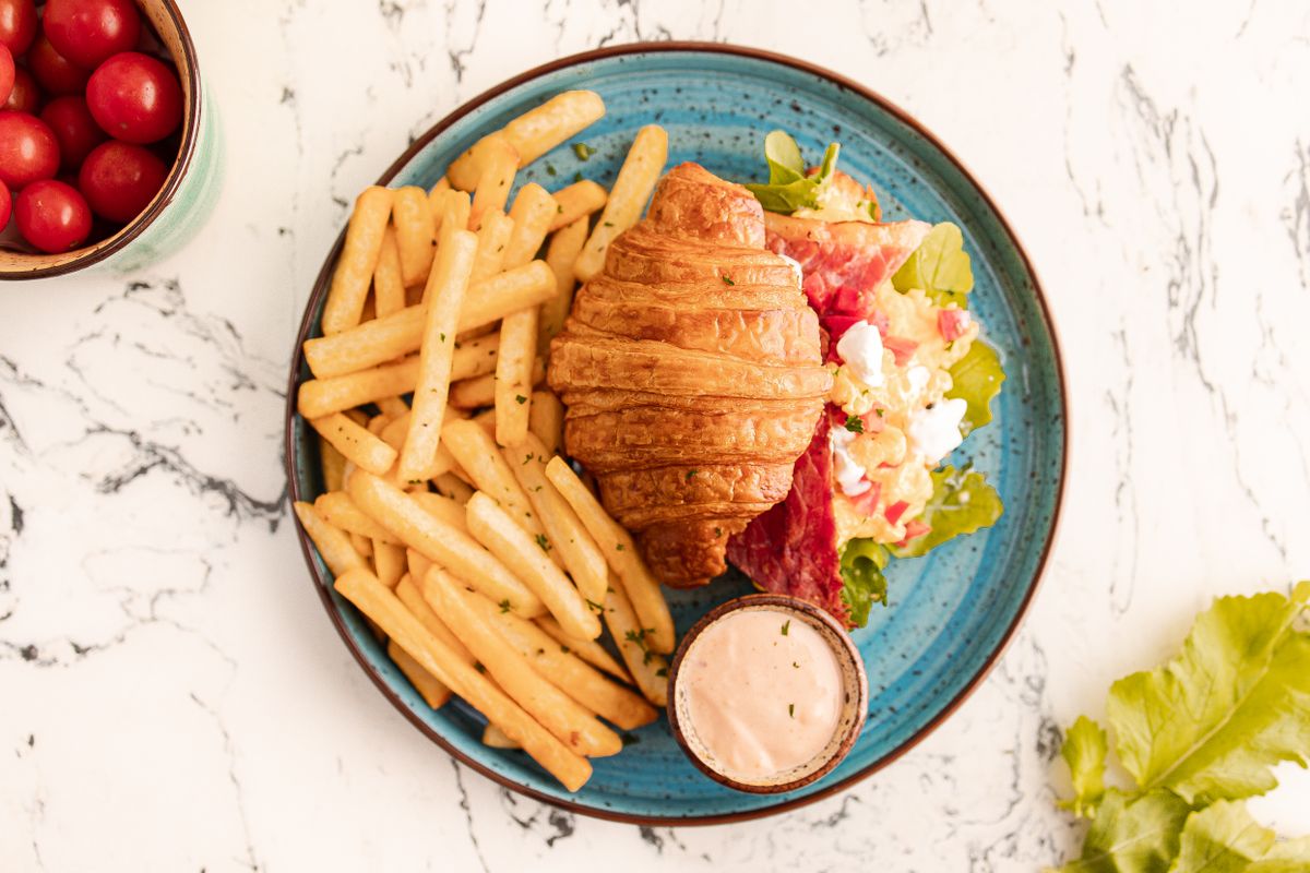 Tasty breakfast featuring a croissant and fries, served with fresh salad and sauce on a rustic plate.