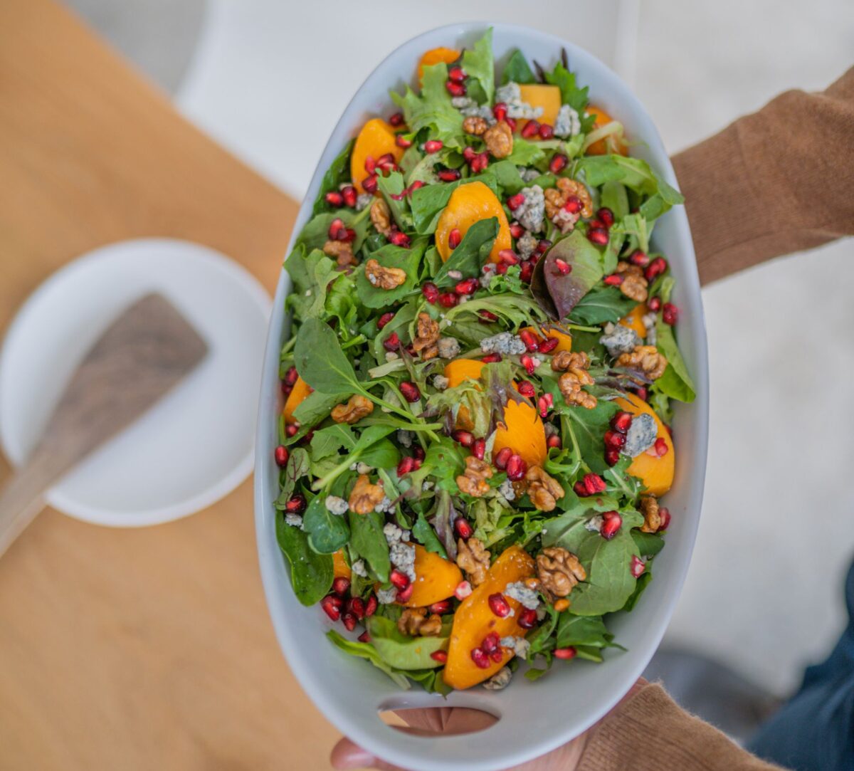 Top view of a vibrant arugula, peach, and walnut salad in a white bowl, held by human hands.