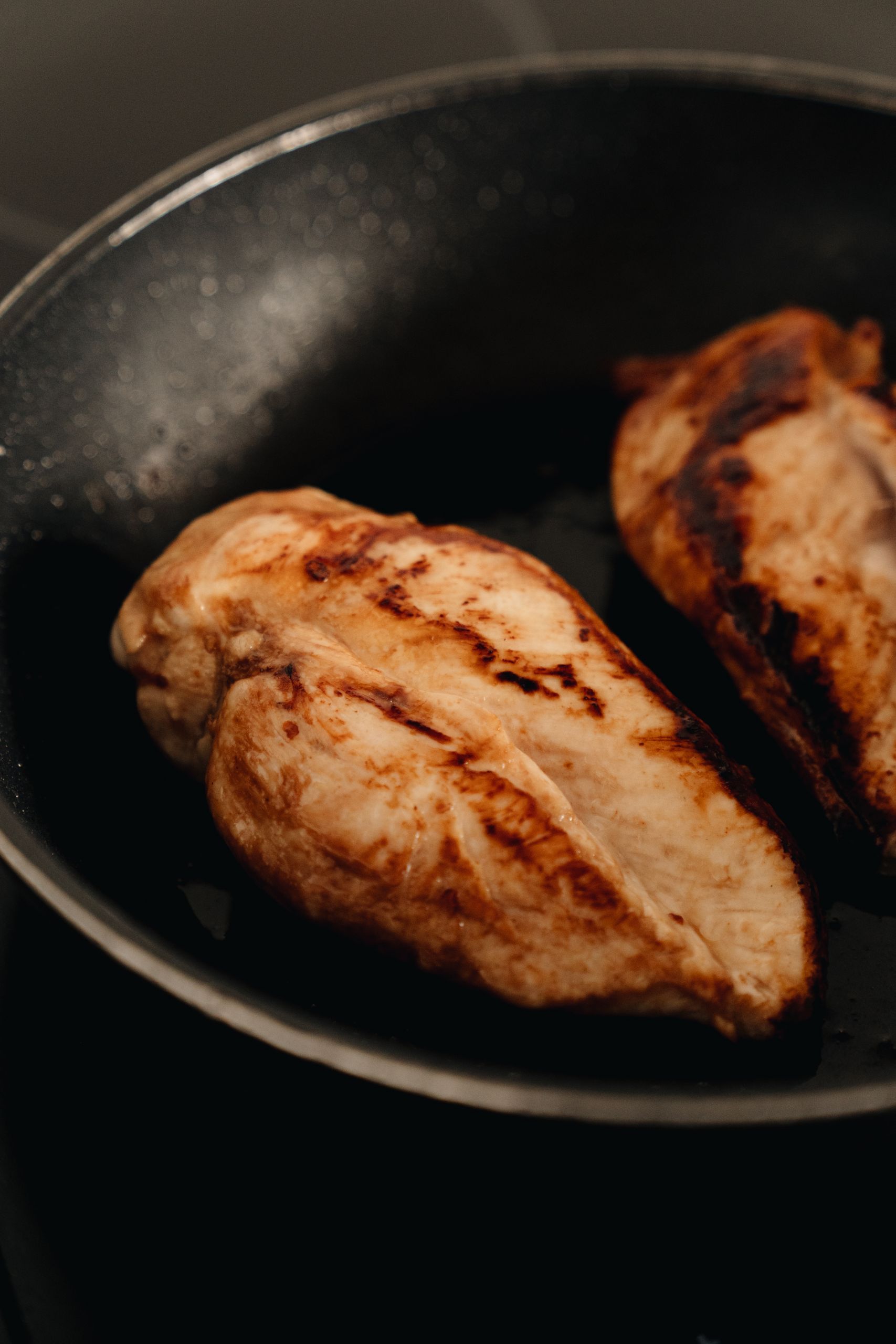 Two beautifully cooked chicken breasts searing on a non-stick frying pan.