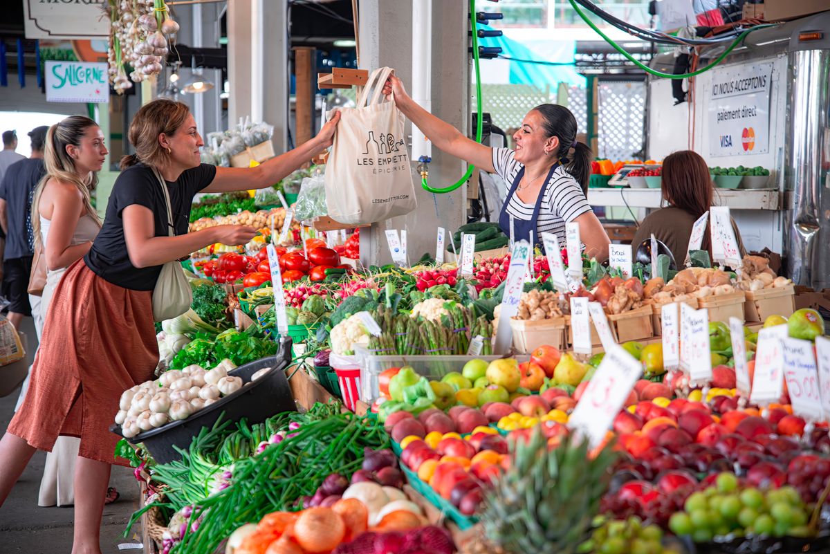 Women shopping at a colorful outdoor market stall filled with fresh fruits and vegetables. Smiles and interaction.