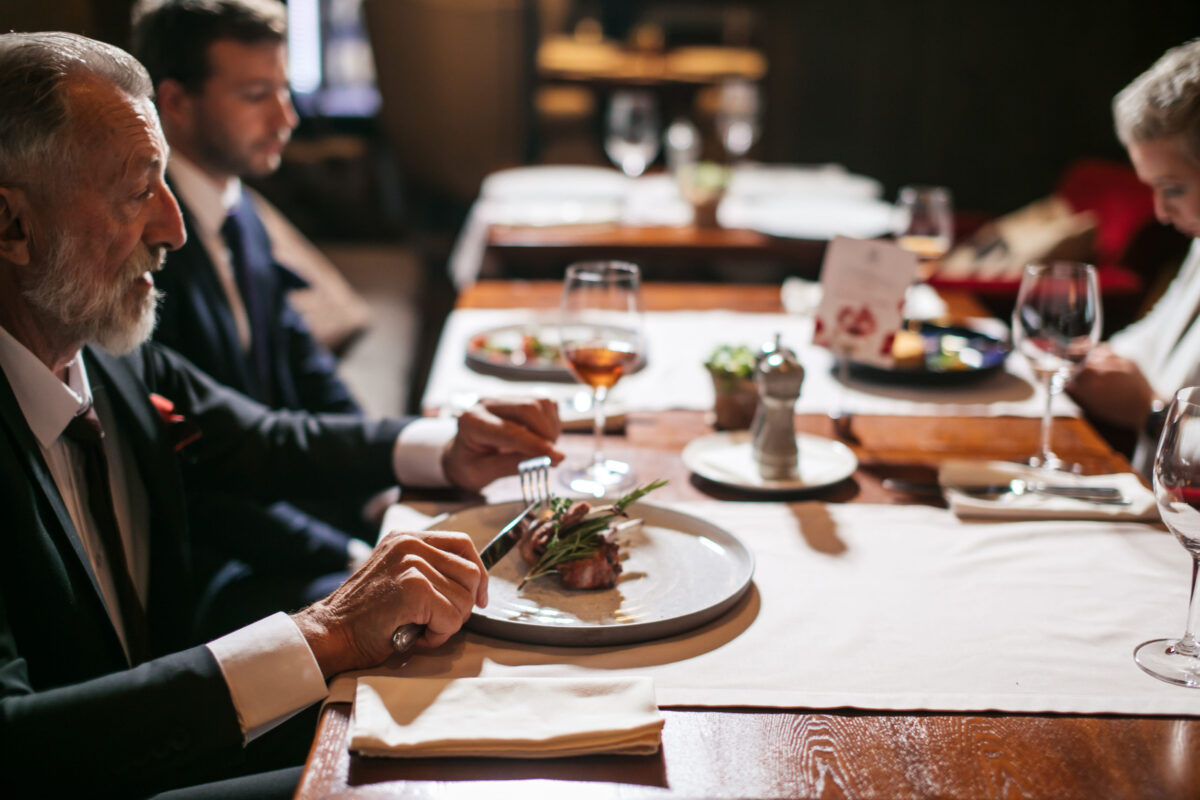 Close up of old bearded man with calm face, holding silver knife and fork ready to start eating delicious meal, in luxury place, horizontal shot
