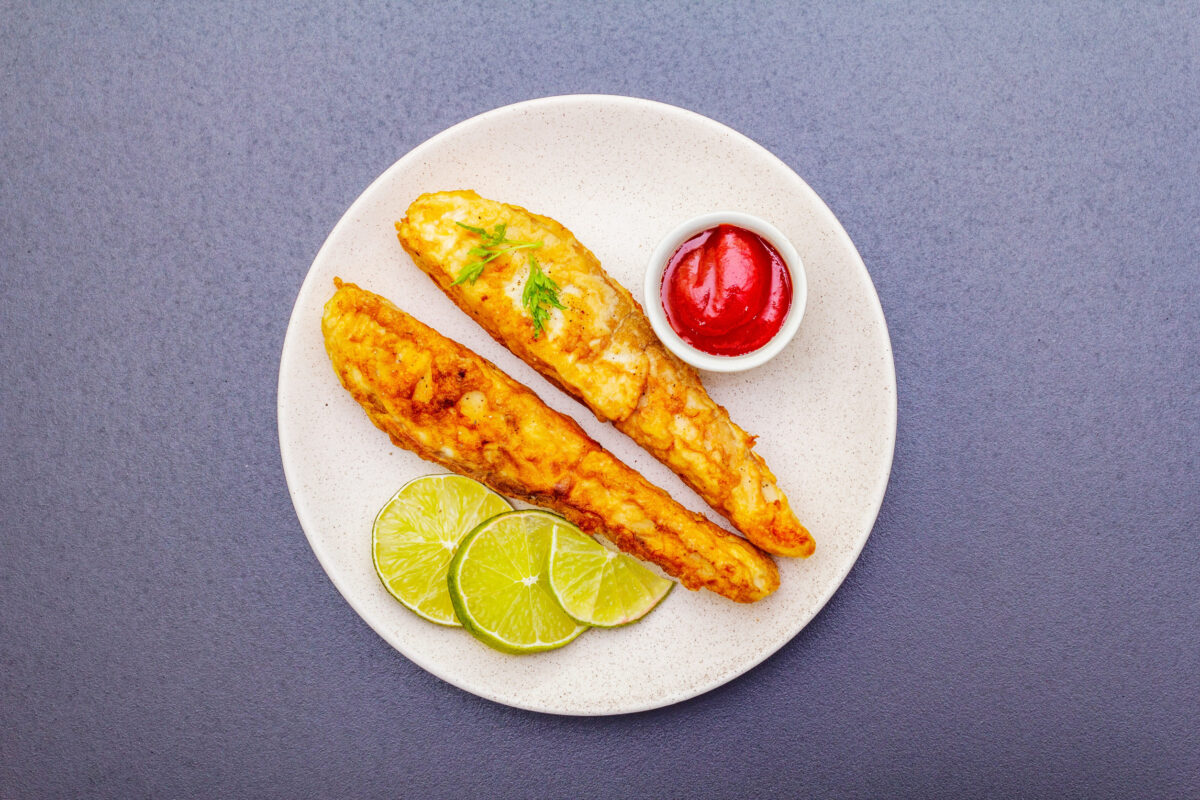 Grilled (fried) pollock in batter. On ceramic plate with lime, parsley and sauce. Cooking fresh fish. On black stone background, top view