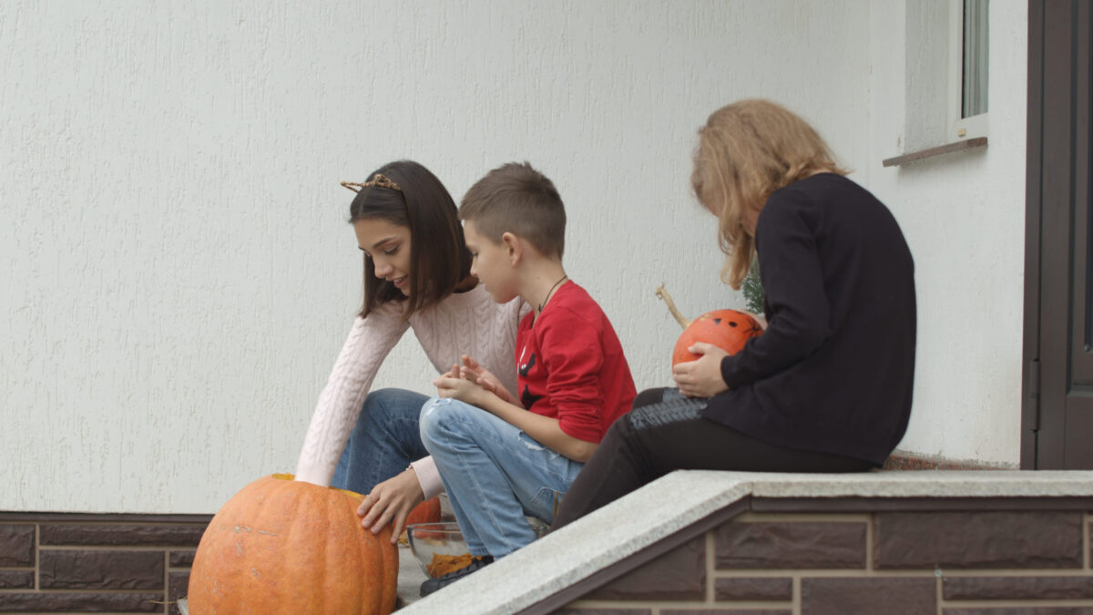 The family is preparing for Halloween on the porch of the house. The boy takes out the insides of the pumpkin. The girl draws a terrible monster.