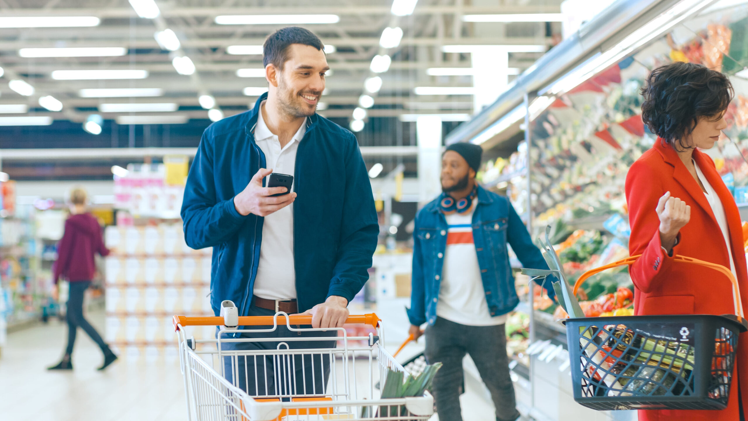 At the Supermarket: Handsome Man with Smartphone, Pushes Shopping Cart, Walks Through Fresh Produce Section of the Store, Chooses Some Products. Other Customers Purchasing Products.