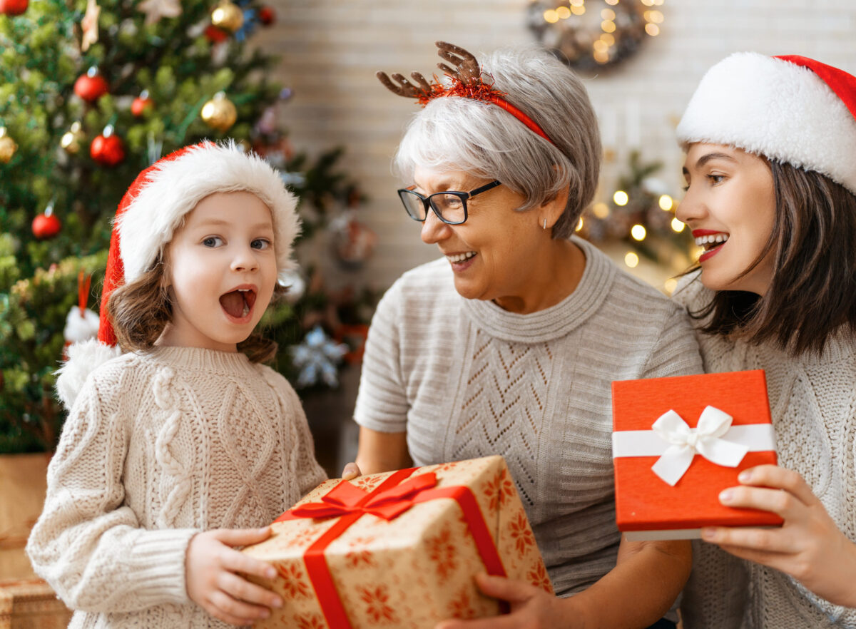 Merry Christmas and Happy Holidays! Cheerful kid presenting gifts to mom and granny. Parents and little child having fun near tree indoors. Loving family with presents in room.