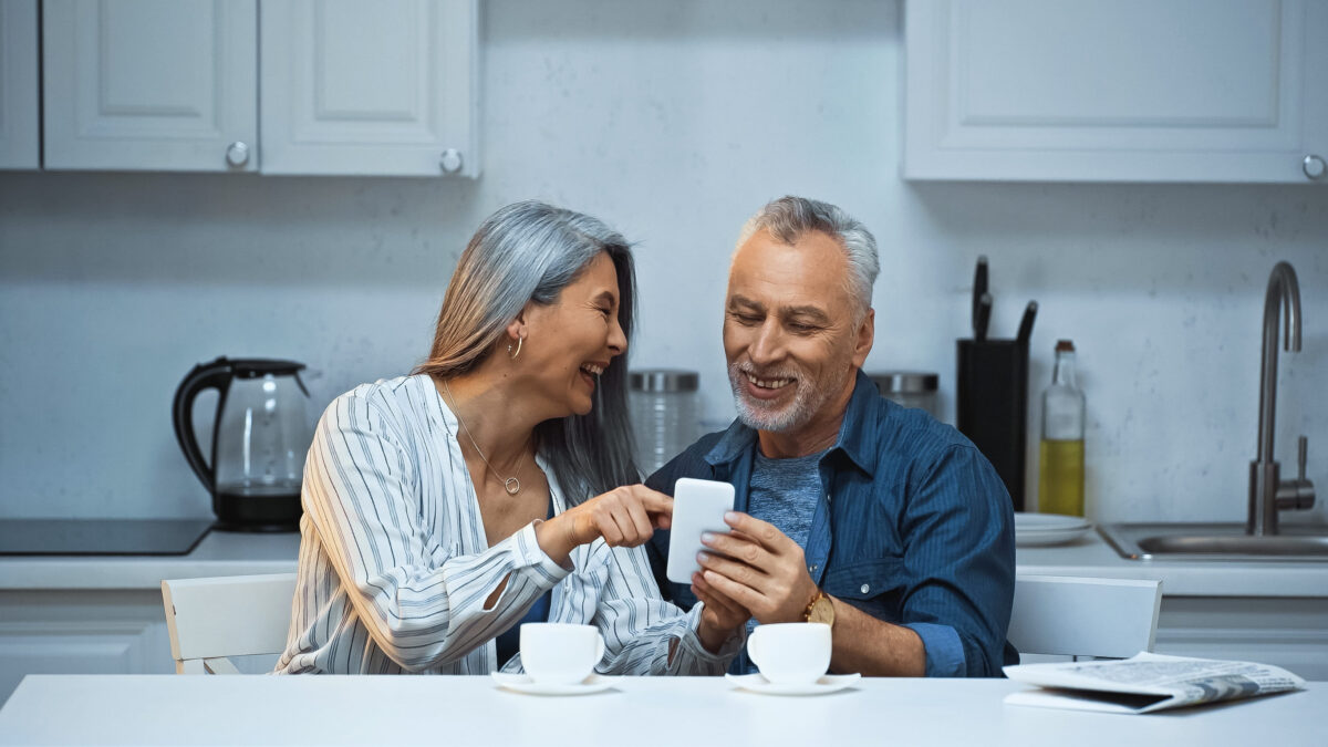 9 Qualities Men May Find More Attractive in a Woman After 50 1 laughing asian woman pointing with finger at smartphone near smiling husband