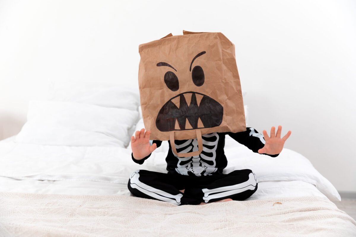 a child in a halloween costume sits with face mask on bed with white bedclothing