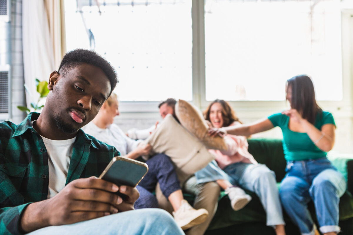 Social isolation problem concept. Selective focus on lonely sad african boy using his smartphone. Blurred background with friends laughing, joking and having fun.