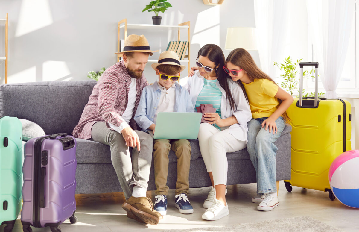 Happy family of four with kids buying tickets and booking hotel online using laptop sitting on sofa in living room at home with packed suitcases planning summer vacation. Holiday trip concept.