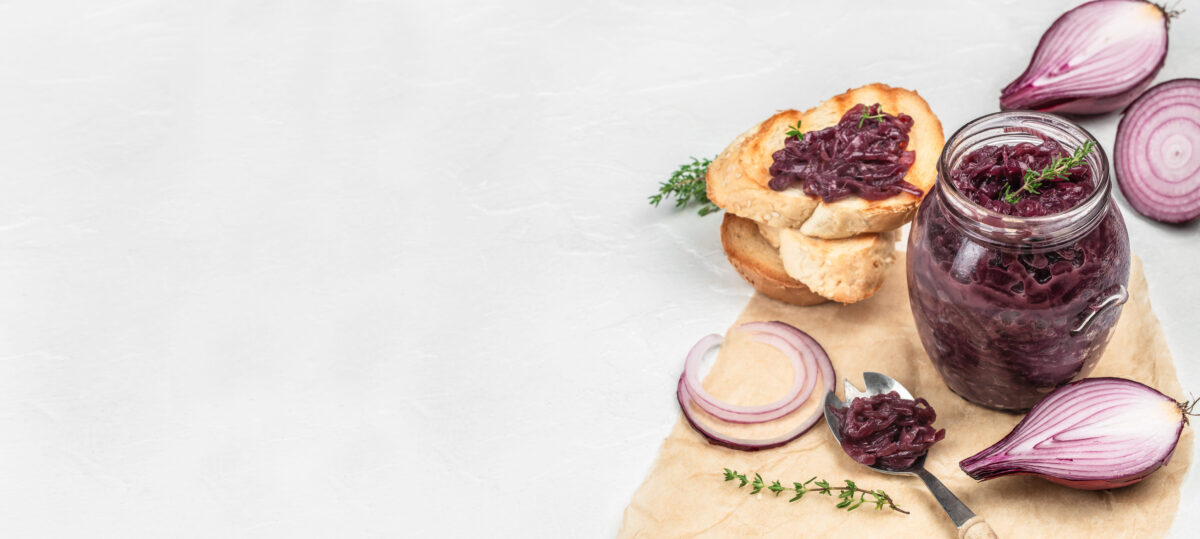 Glass jar of homemade caramelized red onion jam with thyme, served with toasted sesame bread slices, fresh red onion halves, and rings, on a white background copy space