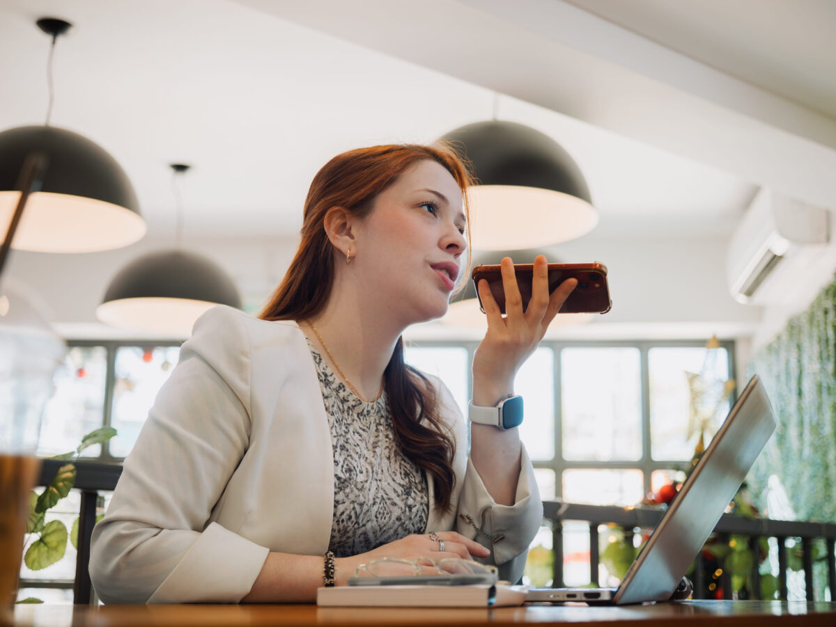 Professional woman holds a smartphone near her mouth while sitting at a laptop in a cafe. Voice messaging and remote work concept.
