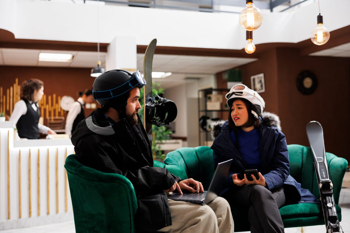 Boyfriend and girlfriend talking and holding their smart devices at cozy lounge of ski resort. Couple in winter clothing with laptop and mobile phone chatting in hotel lobby. Snow gear on display.