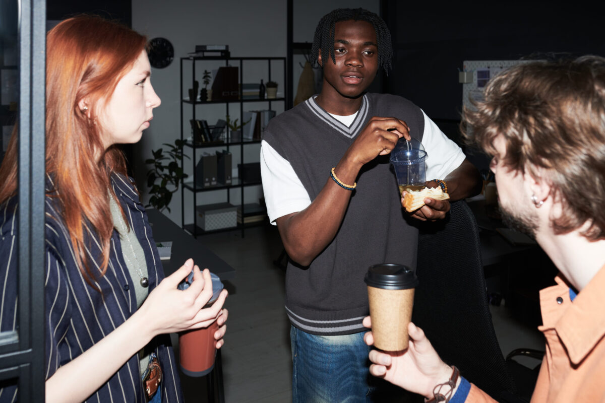 Young adult coworkers discussing project while drinking coffee during office beverages, representing team communication and collaboration. Useful for workplace culture or startup marketing