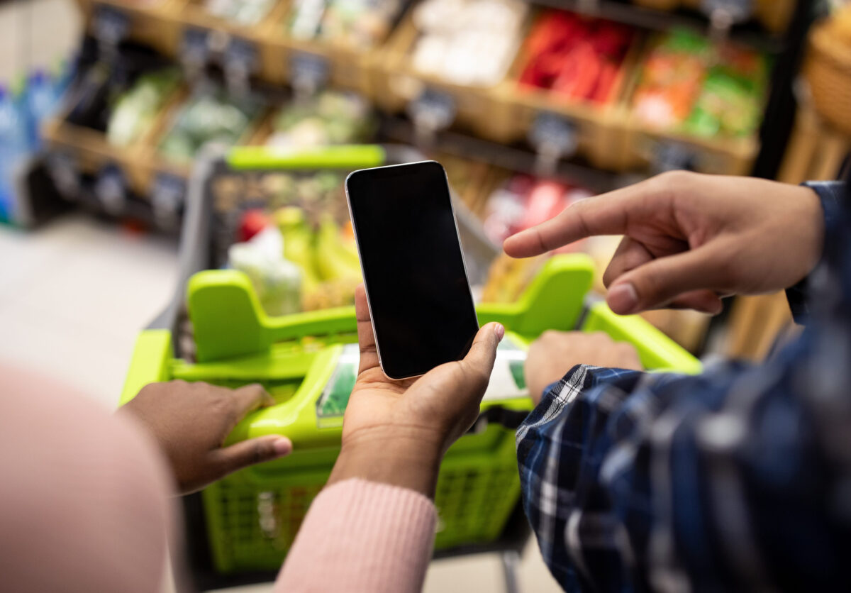 Unrecognizable black couple holding smartphone with empty screen at supermarket, mockup for your mobile app or website. African American guy and lady with cellphone shopping at mall