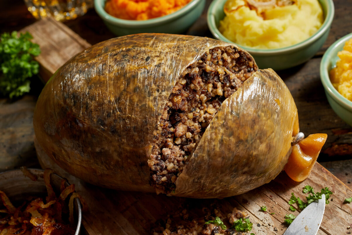 Close up on a sliced open cooked Scottish haggis on a cutting board with side dishes of potato, turnips and carrot, or neeps and tatties