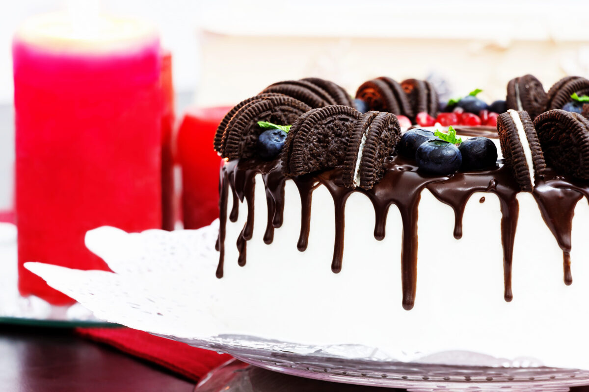 White cream cake with chocolate stains, fresh berry blueberry and pomegranate and chocolate round cookies. Red candle backgroud. Romantic concept. Selective focus.