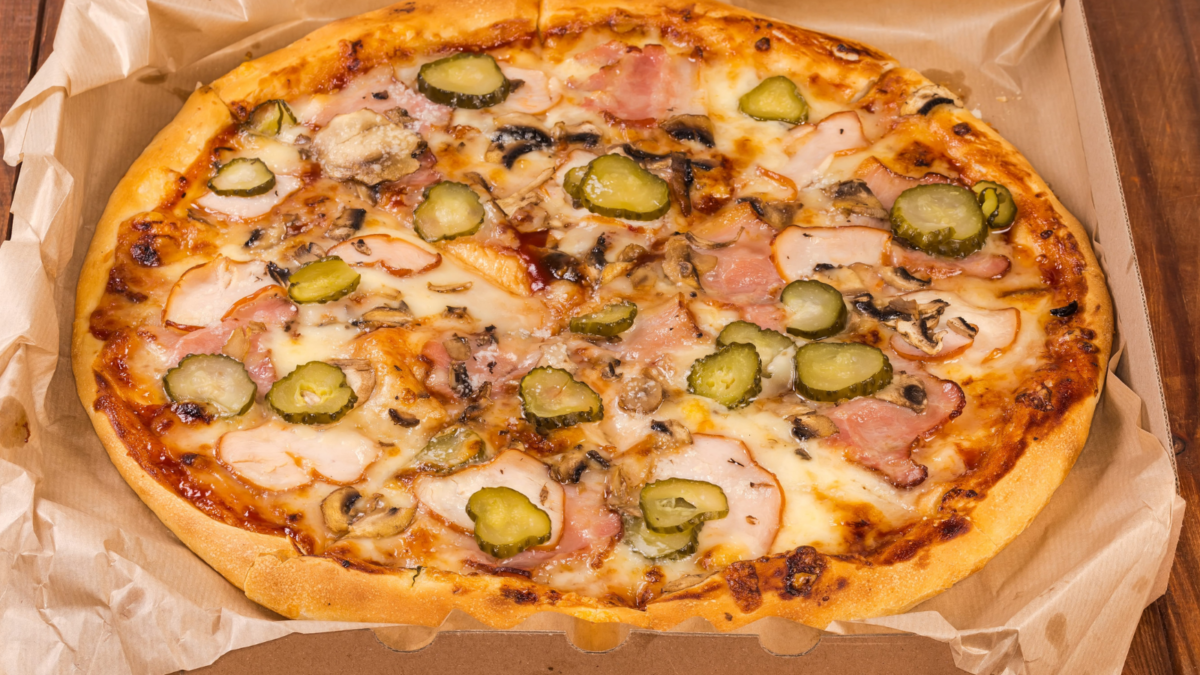 A delicious pizza topped with savory ham, pickles, and melted cheese, resting in a rustic cardboard box, ready to satisfy appetites on a cozy evening. Image Credit: anmbph/ 123RF