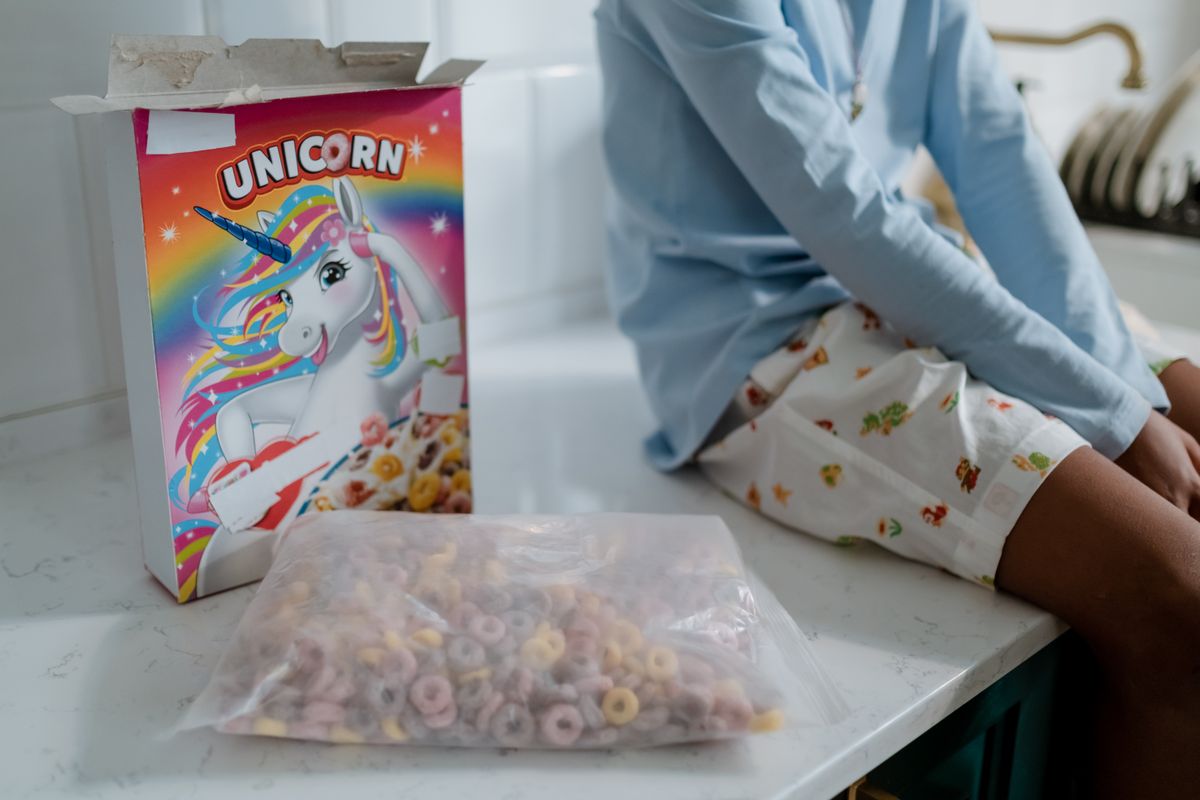 A child sitting on a kitchen counter with an open box of unicorn-themed cereal, highlighting a playful morning scene.