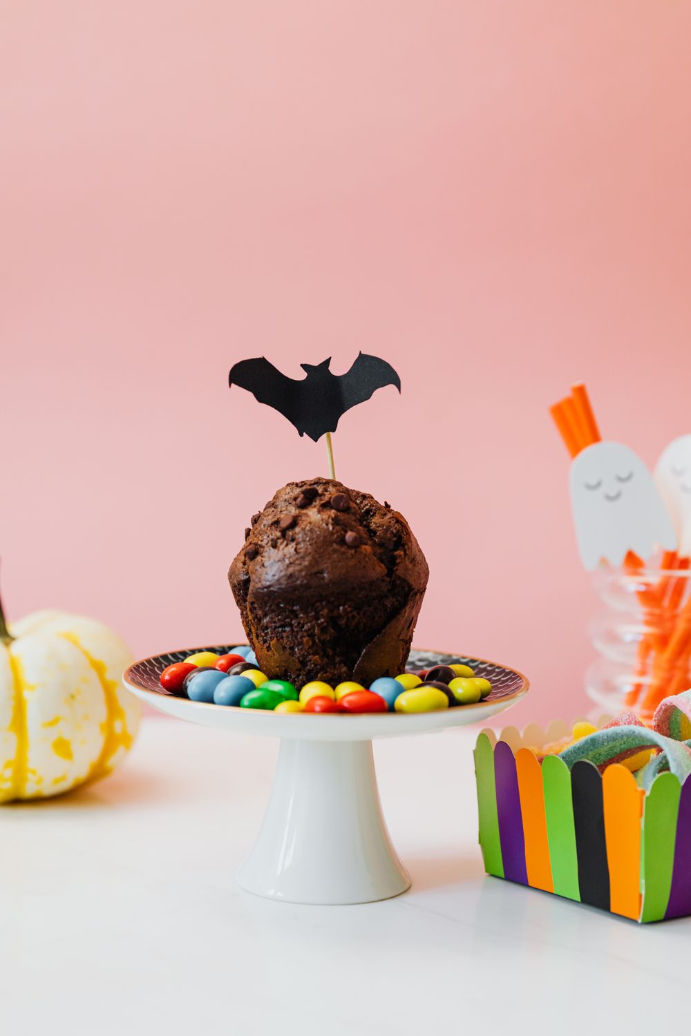 A chocolate muffin with a bat topper surrounded by candies on a Halloween-themed setup.