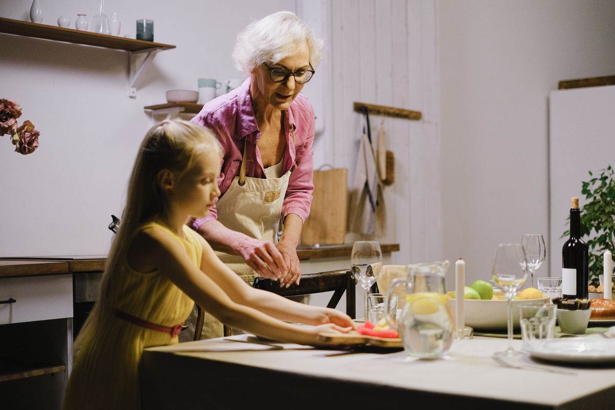 8 Things Grandma Never Threw Away in the Kitchen 1 A grandmother and young granddaughter setting a dining table together indoors.