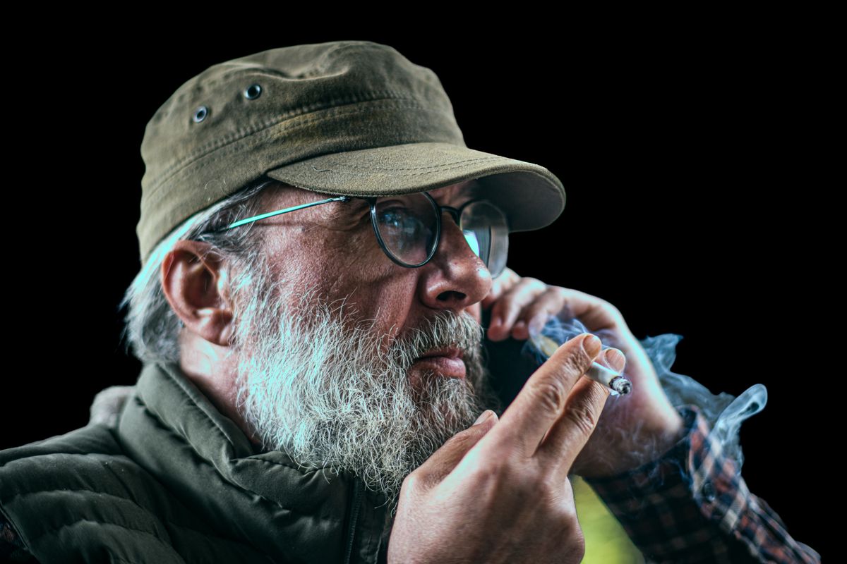 A mature man with a beard smoking and talking on the phone, wearing a cap and glasses.