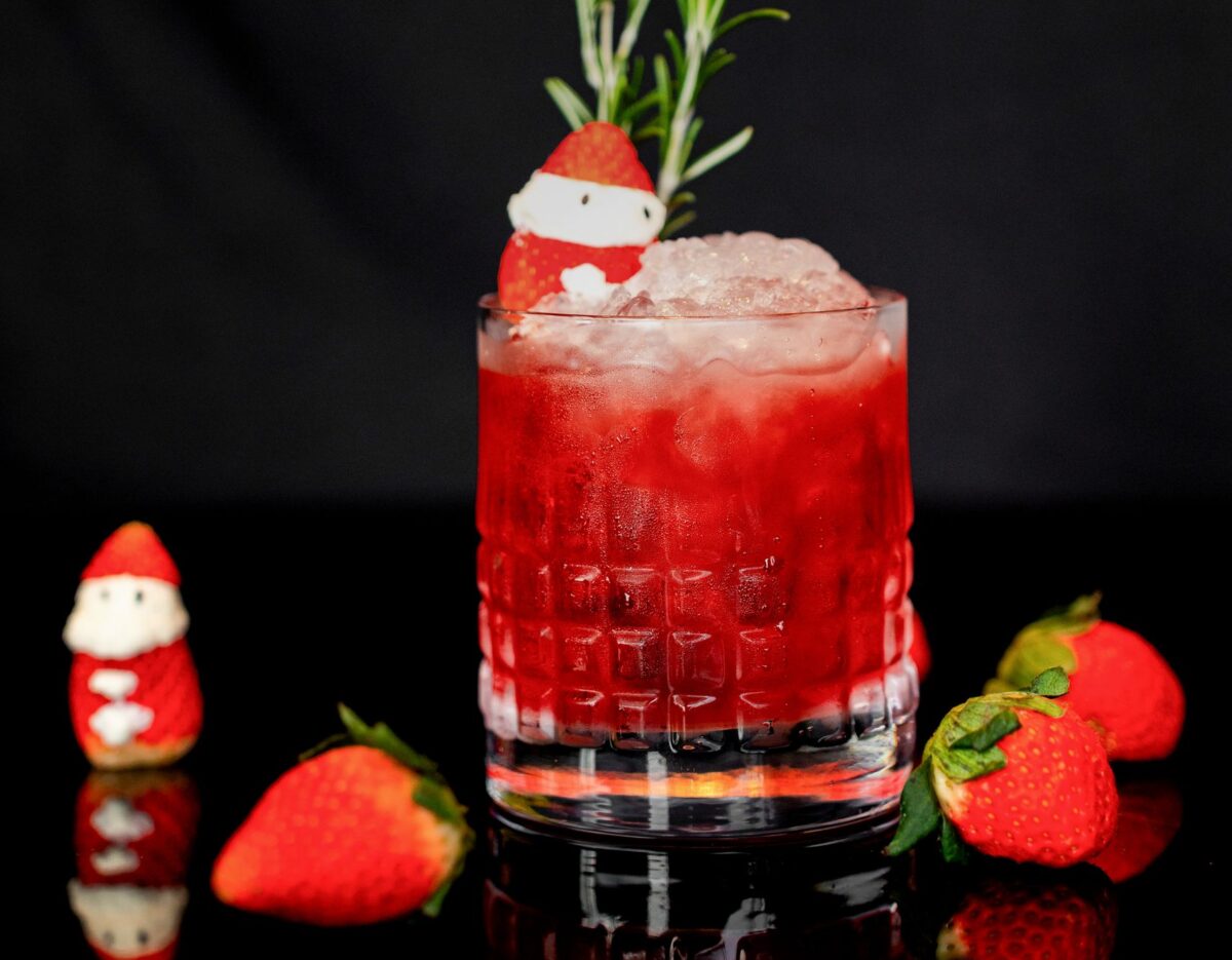 A refreshing strawberry cocktail with rosemary, ice, and festive decorations on black background.