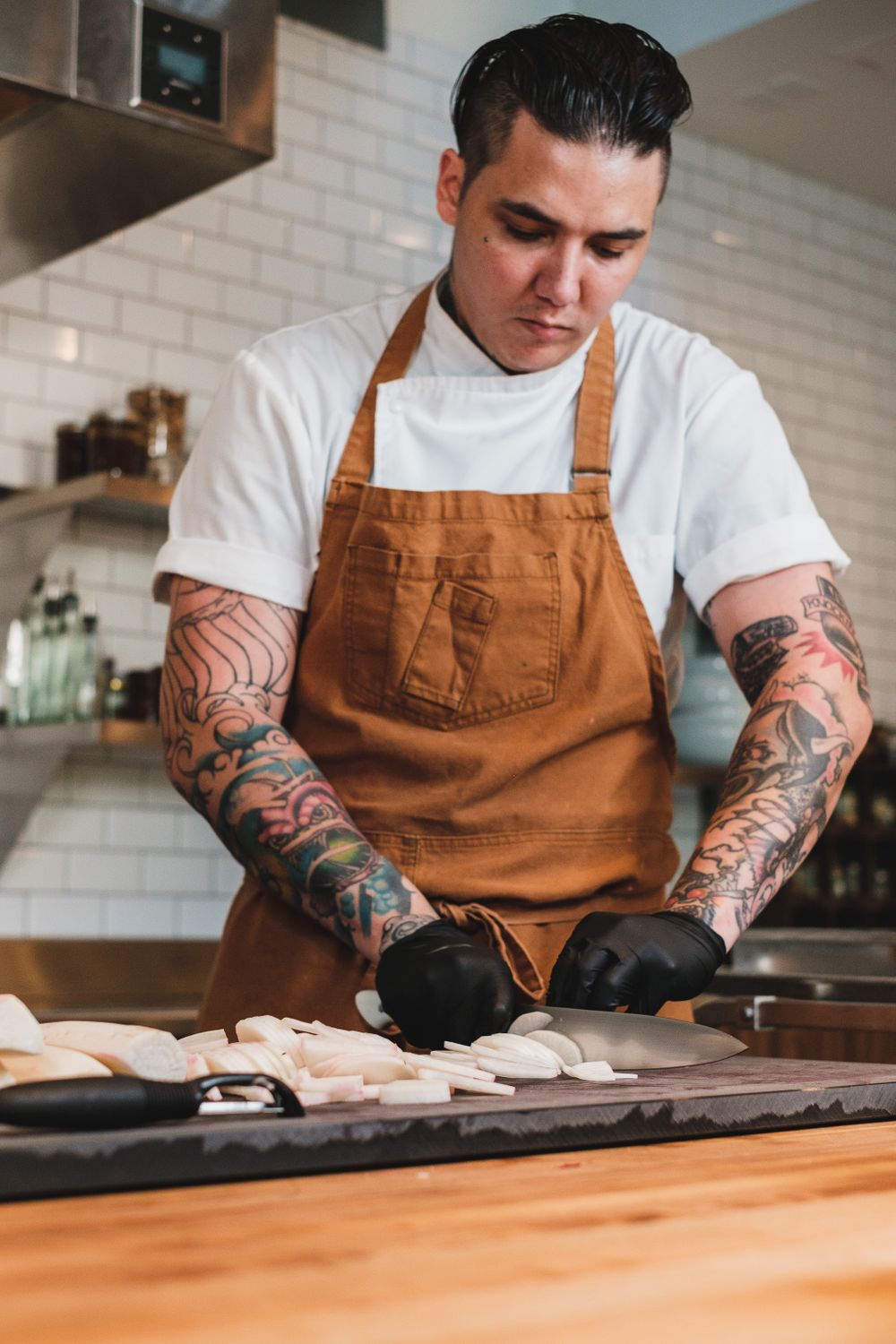 A tattooed chef skillfully slicing ingredients on a wooden board in a professional kitchen setting.
