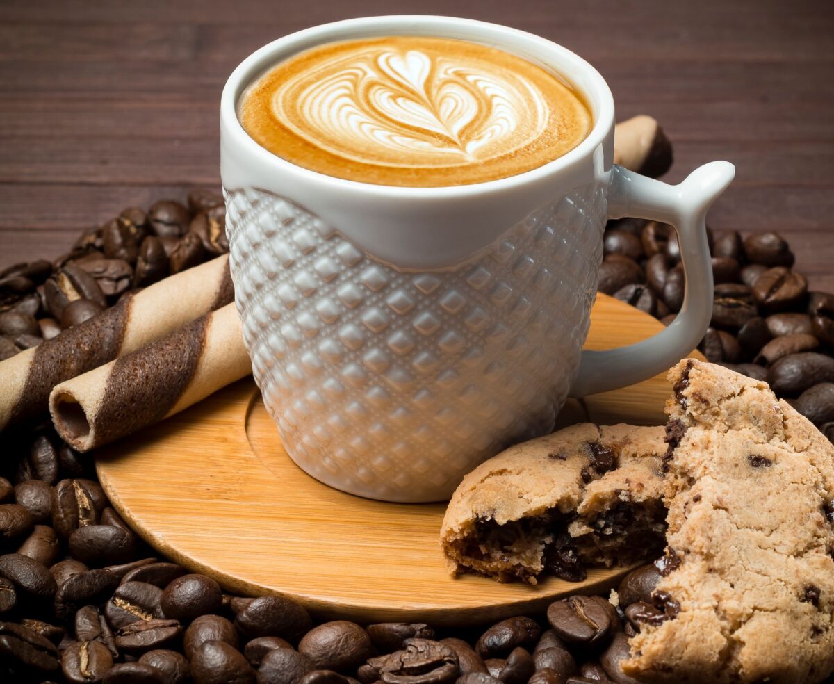 A top view of a coffee cup with latte art, surrounded by coffee beans, a cookie, and a wafer stick.